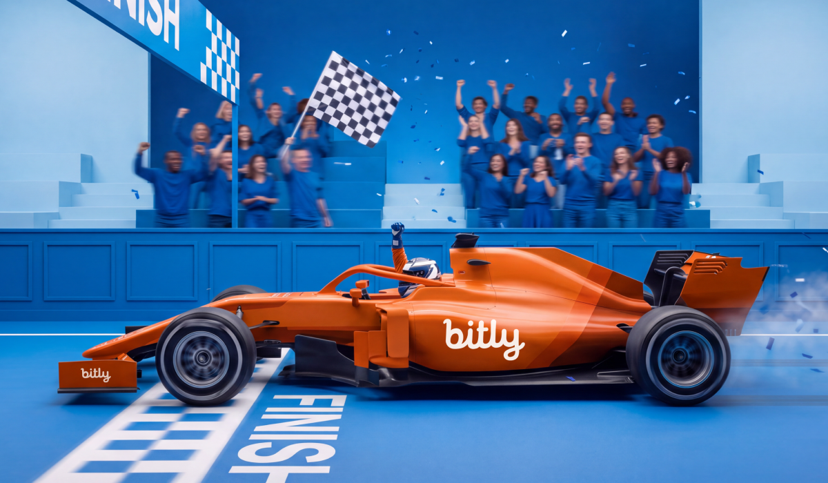 An orange Bitly race car crosses the finish line as a checkered flag waves and a crowd dressed in blue cheers.